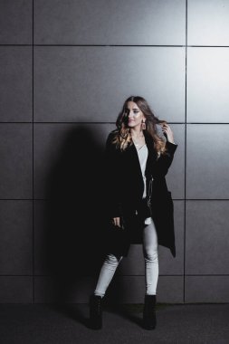 Akşamları karanlık bir duvara karşı poz veren seksi kız. Uzun siyah ceketli, beyaz kotlu, deri çantalı şık bir kız binanın duvarının yanında duruyor. Yürüyüşe çıkan güzel kız.