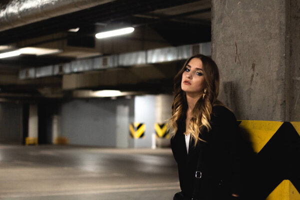 Attractive young brown-haired woman posing in an underground parking. Girl model in black coat posing in an empty underground parking. Fashionable photo of young beautiful girl with evening make-up