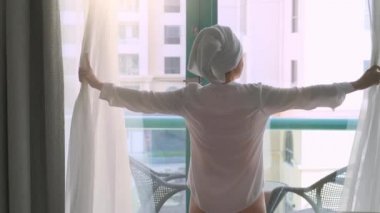 Duştan sonra kafasında havlu olan kadın otel penceresinin perdelerini açıyor.