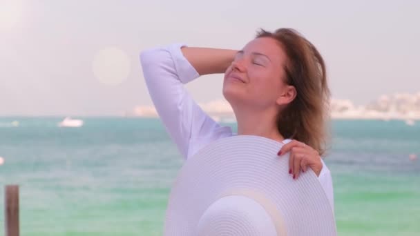 Jonge vrouw in een wit shirt staat voor de zee en straalt haar — Stockvideo