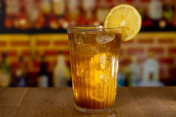 Glass with sparkling cola, ice and lemon on a wooden counter in front of a bar — Stock Photo, Image