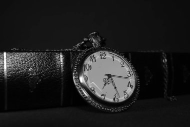 Eski cep saati siyah deri kitaba yaslanmış, siyah beyaz fotoğraflanmış.
