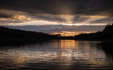 Bir orman gölünde gün batımı sahnesi. Güneş ışınları kara bulutları delip geçiyor.