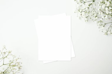 Beyaz masadaki beyaz zarflı boş tebrik kartı modeli. Bebek nefesli çiçek çerçevesi. Üst görünüm, düz döşeme, beyaz kopya alanı. Basit ve minimalist bir kavram..