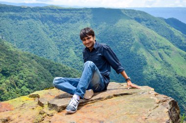 Dağın köşesinde oturan genç adam kameraya poz veriyor. Dağdan turist manzarası. Genç çocuk dağın tepesinden doğanın tadını çıkarıyor..