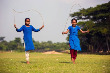 Gowainghat, Bangladeş - 06 Kasım 2019: Çocuklar veya çocuklar açık yeşil çimenler üzerinde ip atlama oyununun keyfini çıkarıyorlar. Okul bahçesinde okul elbiseleriyle ip atlama oyunu oynayan genç kızlar..