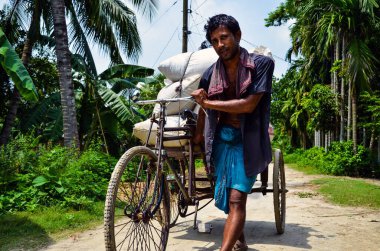 Pabna, Bangladeş - 25 Temmuz 2012: Bir triportör aracının kamyonetteki yükü taşımak için çok çalışması. Zavallı genç adam yaz sıcağında çamurlu bir yolda ağır yüklü metal minibüsü çekiyor..