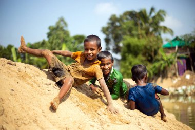 Taherpur, Bangladeş - 05 Kasım 2019: Çocuklar kum tepesinden nehir suyuna yuvarlanıyor. Çocuklar sel sırasında okul tatilinin tadını çıkarıyorlar. Nehir kenarında oynayan ve devrilen çocuklar.
