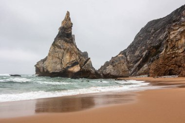 Yüksek kayalıkları ve turkuaz suyu olan kumlu bir sahil manzarası.
