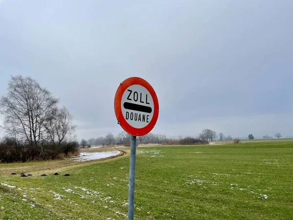 Silniční značka označující celnice, Zoll, douane, mezi rakouskou a lichtenštejnskou hranicí, v zelené krajině proti modré obloze. — Stock fotografie