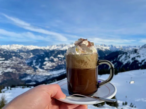 Apres esqui, depois de esqui nos Alpes austríacos. Mão segurando xícara de chocolate quente com chantilly, montanhas cobertas de neve no fundo. — Fotografia de Stock