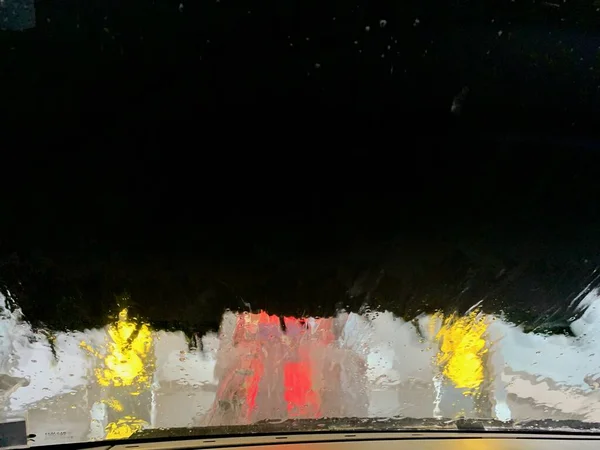 Inside automatic conveyorized tunnel car wash. View from inside car, windshield with water drops and soap foam, black rotating brushes. — Stock Photo, Image