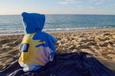 Kapüşonlu havluya sarılı çocuk kumlu sahilde oturmuş denize bakıyor..