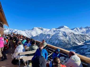 Lech, Avusturya, 23.01.2022. Güneşli bir kış gününde, Arlberg kayak sahasının bir parçası olan Lech 'teki dağların muhteşem manzarasıyla dağ restoranının terasında dinlenen insanlar..