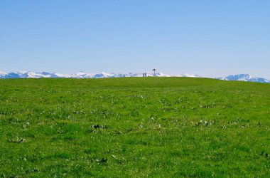 Mavi gökyüzüne karşı yeşil çayır, millet, yol kesişmesi ve ufukta karlı dağ sıraları. Hochberg, Pfaender yakınlarında. Vorarlberg, Avusturya.