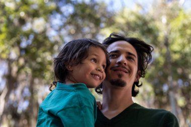 Latin Amerikalı baba 3 yaşındaki oğlunu parkta kollarında taşıyor. Yakın çekim. Aile ve Babalar Günü konsepti.
