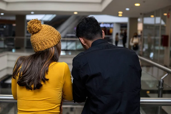 İki Latin Amerikalı sırtı alışveriş merkezinin parmaklıklarına dönük duruyor. Kadın sarı bir şapka ve uzun kollu fanila giyiyor, adam da siyah. Çift kavramı