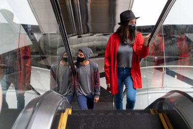 Yüz maskeli iki genç Latin Amerikalı kardeş alışveriş merkezinde yürüyen merdivenlere tırmanıyor. Yürüyüş ve yüz maskesi kavramı