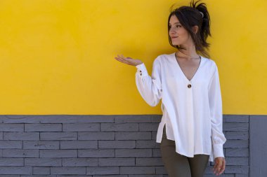 Gülümseyen genç Latin Amerikalı kadın portresi (24), sarı ve gri arka planda izole edilmiş, avuç içi boşluğu işaret ediyor. Tanıtım indirimi teklifini kabul et.