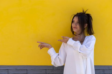 Gülümseyen genç Latin Amerikalı kadın portresi (24) sarı ve gri arkaplan noktalarında, elinin iki parmağıyla boş uzaya izole edilmiştir. Tanıtım indirimi teklifini kabul et.