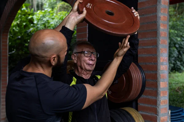 Yaşlı bir Latin Amerikalı adam ve antrenörü ellerinde spor salonunda rehabilitasyon ve terapi için kullandıkları bir ağırlık diski tutuyorlar. Sağlıklı yaşam tarzı kavramı.