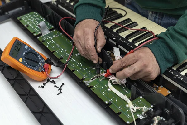 Hands of Latin American electronic technician with a multimeter makes current and resistance measurements to an electronic board of an electric piano in a service center. Repair concept, piano, electronics