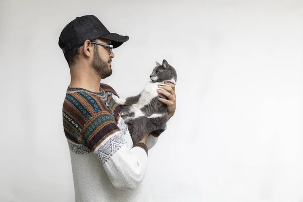 Latin American man standing in profile with cap and glasses looks straight ahead at his cat while holding her in his arms. White background. Pets Concept