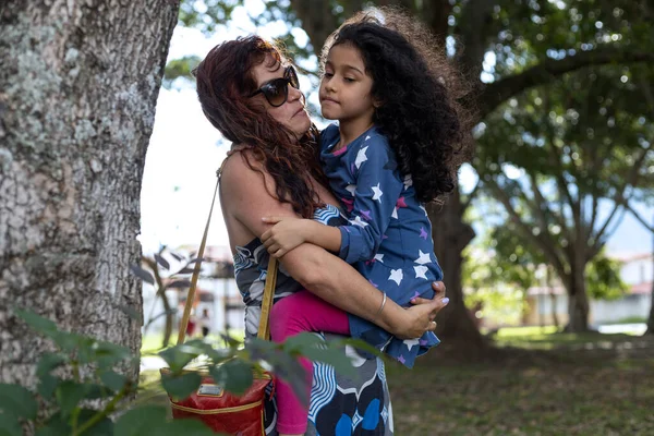 Mom and her Latina daughter share together in the park, she holds her daughter in her arms lovingly