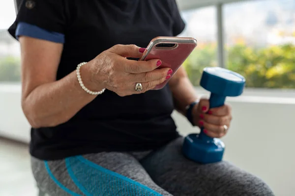 Older Latina woman works out at gym with dumbbells using cell phone workout videos