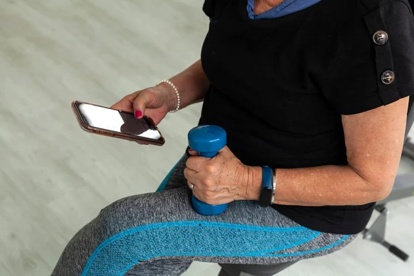 Older Latina woman works out at gym with dumbbells using cell phone workout videos