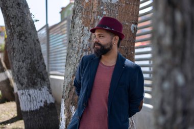 Şehirdeki bazı ağaçların yanında sakallı, şapkalı ve ceketli Latin Amerikalı bohem adamın orta boy fotoğrafı. Tatil konsepti ve portre