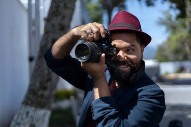 Sakallı, şapkalı ve elinde fotoğraf makinesi olan Latin Amerikalı bohem adam şehirde fotoğraf çekiyor. Kariyer ve tatil konsepti