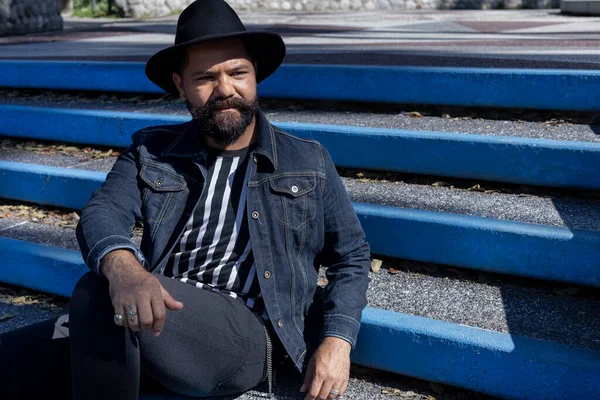 Sakallı ve şapkalı Latin Amerikalı yetişkin bir adam meydanın merdivenlerinde rahatça oturuyor. Hipster ve Tatil Konsepti