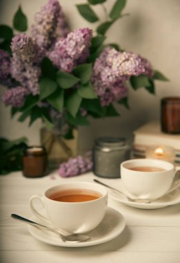 Bir fincan yeşil çay, arka planda bir demet leylağın üzerine serpiştirilmiş gri bir arkaplan. Kitaplar ve mumlarla romantik kompozisyon. Bahar çayı içeceği. Yan görüş. Kopyalanacak yer.