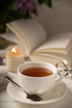 Bir fincan yeşil çay, arka planda bir demet leylağın üzerine serpiştirilmiş gri bir arkaplan. Kitaplar ve mumlarla romantik kompozisyon. Bahar çayı içeceği. Yan görüş. Kopyalanacak yer.