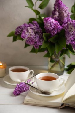 Bir fincan yeşil çay, arka planda bir demet leylağın üzerine serpiştirilmiş gri bir arkaplan. Kitaplar ve mumlarla romantik kompozisyon. Bahar çayı içeceği. Yan görüş. Kopyalanacak yer.
