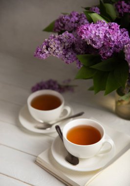 Bir fincan yeşil çay, arka planda bir demet leylağın üzerine serpiştirilmiş gri bir arkaplan. Kitaplar ve mumlarla romantik kompozisyon. Bahar çayı içeceği. Yan görüş. Kopyalanacak yer.