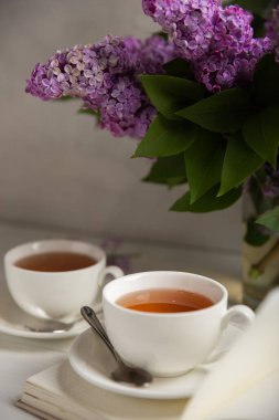 Bir fincan yeşil çay, arka planda bir demet leylağın üzerine serpiştirilmiş gri bir arkaplan. Kitaplar ve mumlarla romantik kompozisyon. Bahar çayı içeceği. Yan görüş. Kopyalanacak yer.