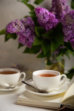 Bir fincan yeşil çay, arka planda bir demet leylağın üzerine serpiştirilmiş gri bir arkaplan. Kitaplar ve mumlarla romantik kompozisyon. Bahar çayı içeceği. Yan görüş. Kopyalanacak yer.