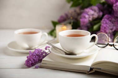 Bir fincan yeşil çay, arka planda bir demet leylağın üzerine serpiştirilmiş gri bir arkaplan. Kitaplar ve mumlarla romantik kompozisyon. Bahar çayı içeceği. Yan görüş. Kopyalanacak yer.