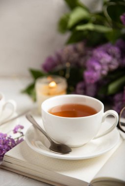 Bir fincan yeşil çay, arka planda bir demet leylağın üzerine serpiştirilmiş gri bir arkaplan. Kitaplar ve mumlarla romantik kompozisyon. Bahar çayı içeceği. Yan görüş. Kopyalanacak yer.