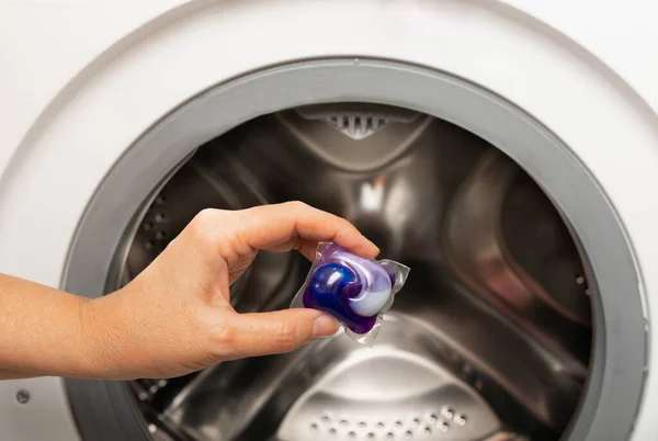 A woman put a liquid powder capsule into a washing machine with laundry, close-up. Gel for washing in the car. Colorful eco-gel for washing in capsules. Washing clothes. Purity concept