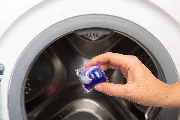 A woman put a liquid powder capsule into a washing machine with laundry, close-up. Gel for washing in the car. Colorful eco-gel for washing in capsules. Washing clothes. Purity concept
