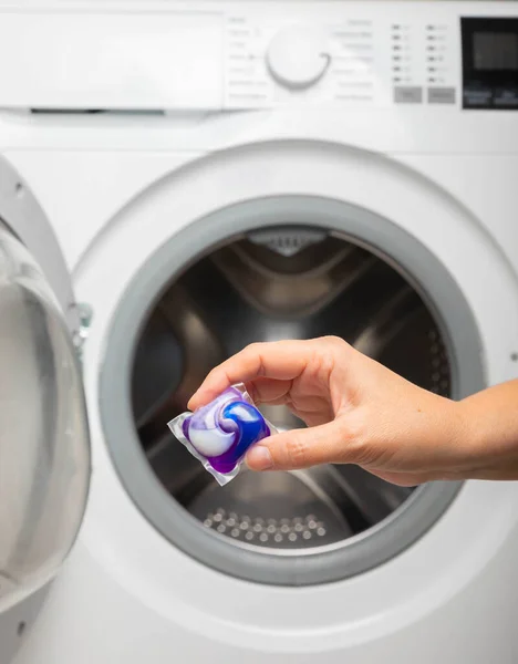 A woman put a liquid powder capsule into a washing machine with laundry, close-up. Gel for washing in the car. Colorful eco-gel for washing in capsules. Washing clothes. Purity concept