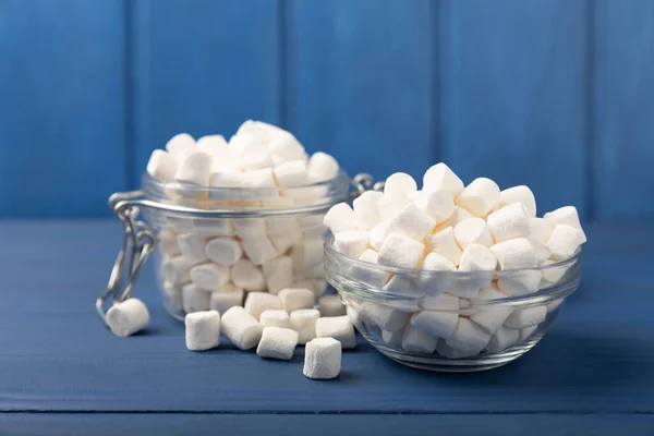 Marshmallow on blue textured wood. White marshmallows in a glass bowl and a jar on the table.Sweets and snacks for a snack. Chewing candies close-up. Copy space. Place for text. Winter food concept.