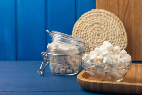 Marshmallow on blue textured wood. White marshmallows in a glass bowl and a jar on the table.Sweets and snacks for a snack. Chewing candies close-up. Copy space. Place for text. Winter food concept.