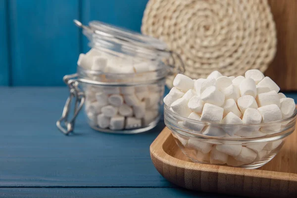 Marshmallow on blue textured wood. White marshmallows in a glass bowl and a jar on the table.Sweets and snacks for a snack. Chewing candies close-up. Copy space. Place for text. Winter food concept.