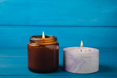 Cozy burning candle in a brown glass jar and a concrete planter on a blue wood texture. winter home decor. Soy ecological candle. Home and interior decoration. Place for text.