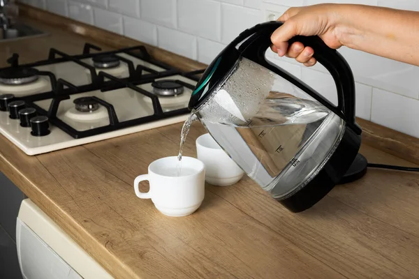 A man makes tea using boiling water from an electric kettle in the kitchen at home. It's time to have breakfast and drink tea. Modern electric kettle on a wooden table. Kettle for boiling water.