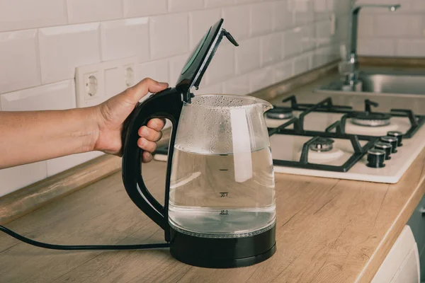 A hand opens the lid of an electric kettle. A modern electric transparent kettle on a wooden table in the kitchen. A kettle for boiling water and making tea. Home appliances for making hot drinks.